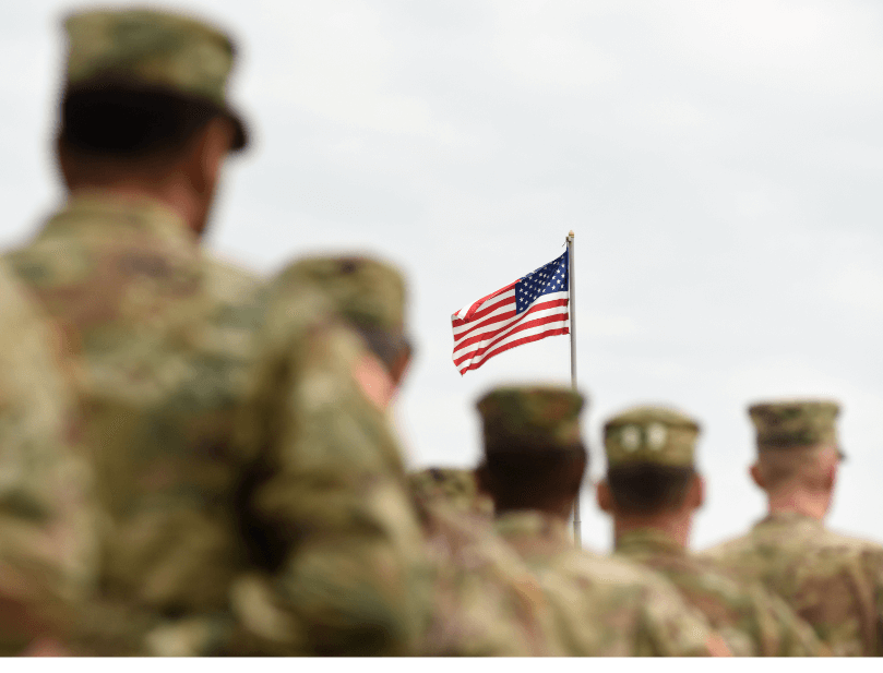 Out-of-focus back view of five military personnel dressed in uniform facing an in-focus American flag