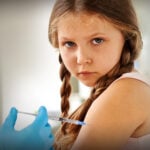 A young girl with a serious look on her face is getting an injection in her arm.