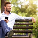 A man in dress clothes is sitting on a park bench smiling with his phone in his hand.