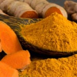 A wooden spoon filled with turmeric powder next to a pile of turmeric.