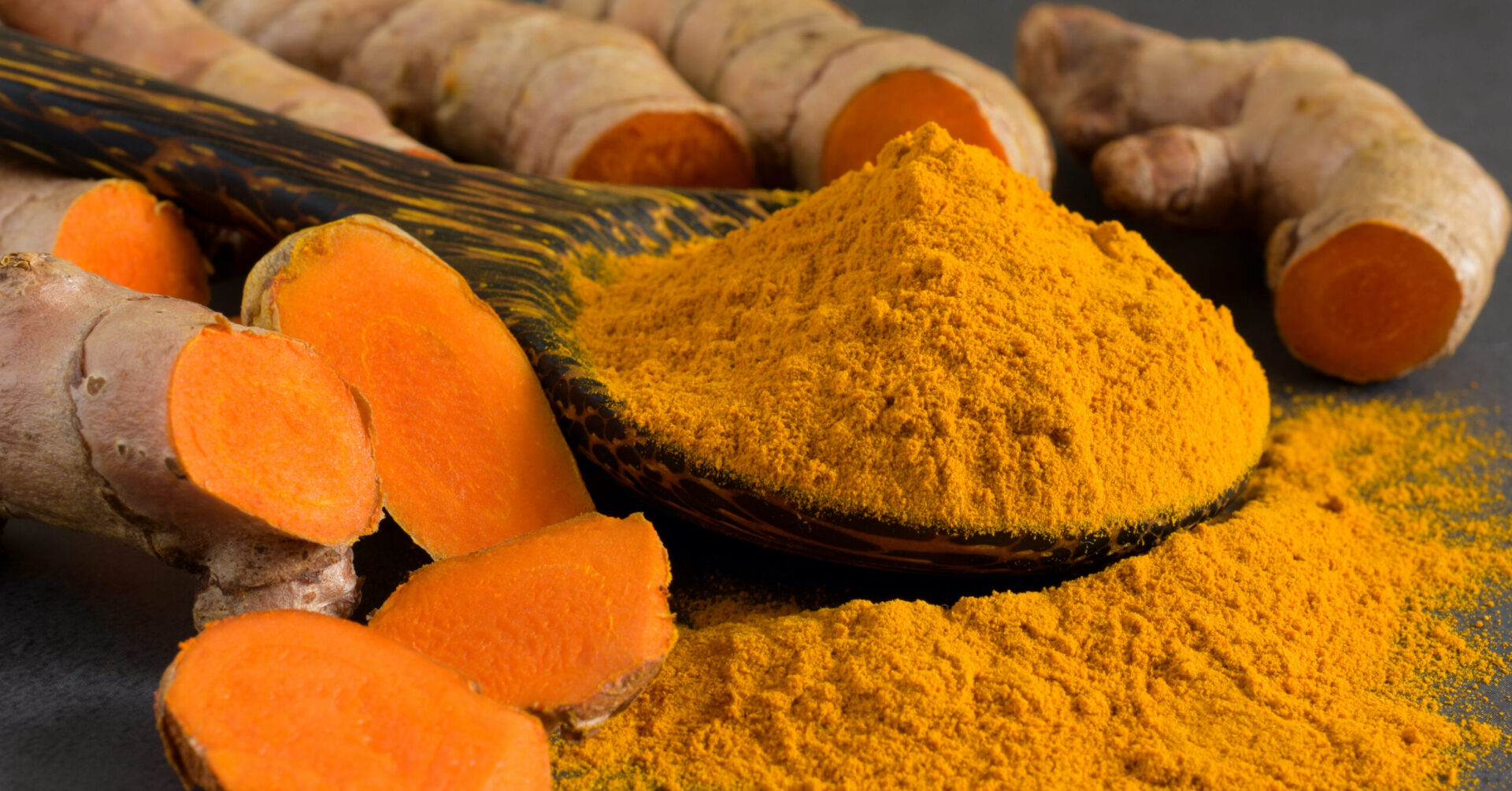 A wooden spoon filled with turmeric powder next to a pile of turmeric.