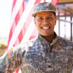 A man in a U.S. Army uniform is smiling in front of an American flag.