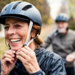 A woman is smiling while putting on a bicycle helmet.