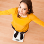 A woman in a yellow shirt is standing on a scale, smiling with her arms outstretched.
